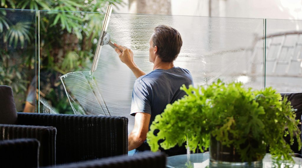 A man cleaning a glass barrier outdoors with a squeegee, surrounded by greenery.